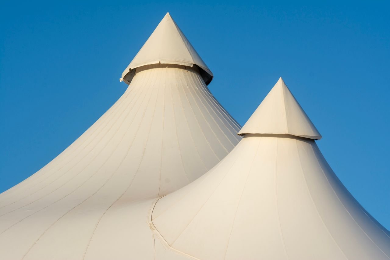 services-02 Two elegant white tent tops with sharp peaks set against a bright blue sky, showcasing modern architecture.